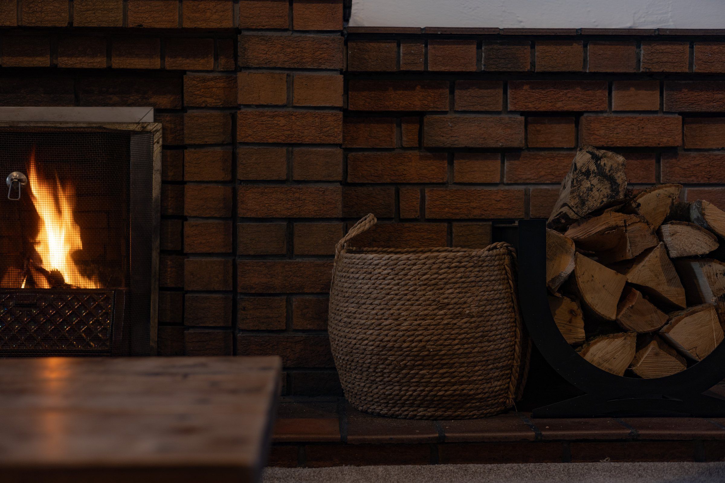 A fire lit in the brick fireplace, with a rope log basket and a stack of logs alongside
